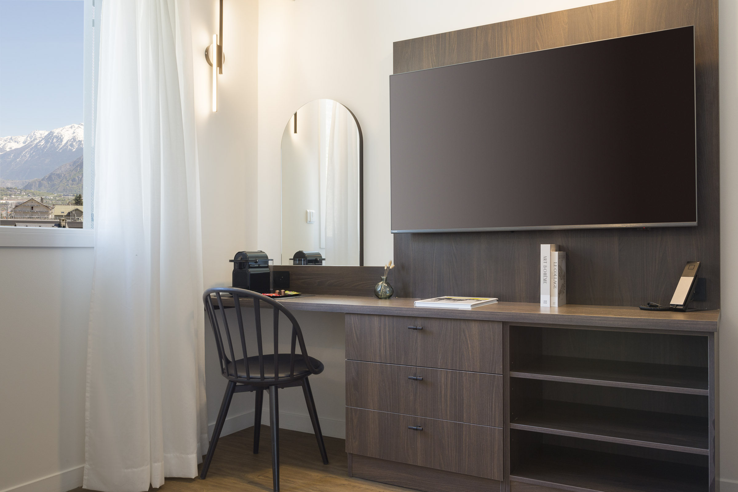 Bureau, télévision murale et fenêtre avec vue montagne dans un appartement 2 pièces du Nehô Suites Saint-Jean-de-Maurienne.