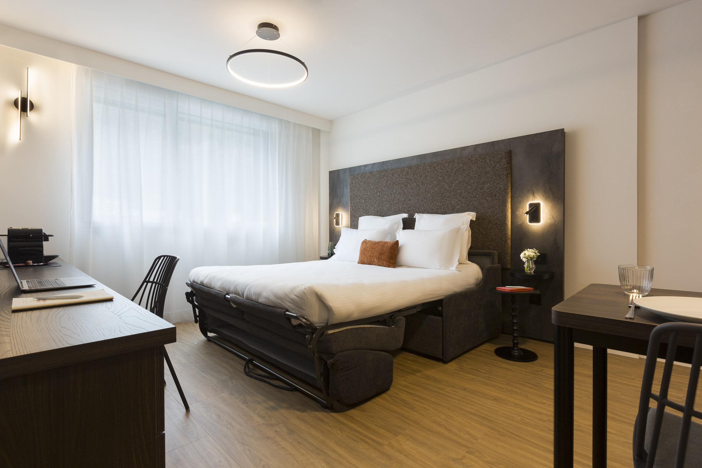 Studio supérieur avec canapé-lit ouvert, bureau, fenêtre et table au Nehô Suites Saint-Jean-de-Maurienne.